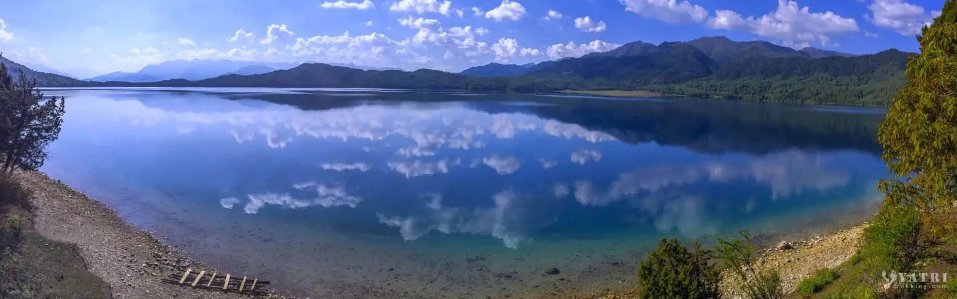 Wide panoramic banner view of crystal-clear Rara Lake reflecting blue skies, clouds, and surrounding green hills in the peaceful landscapes of western Nepal