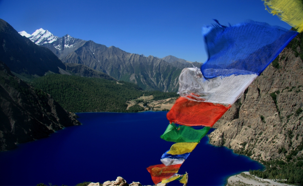 Colorful Tibetan prayer flags fluttering above the deep blue waters of Shey Phoksundo Lake surrounded by dramatic Himalayan mountains in Upper Dolpo.
