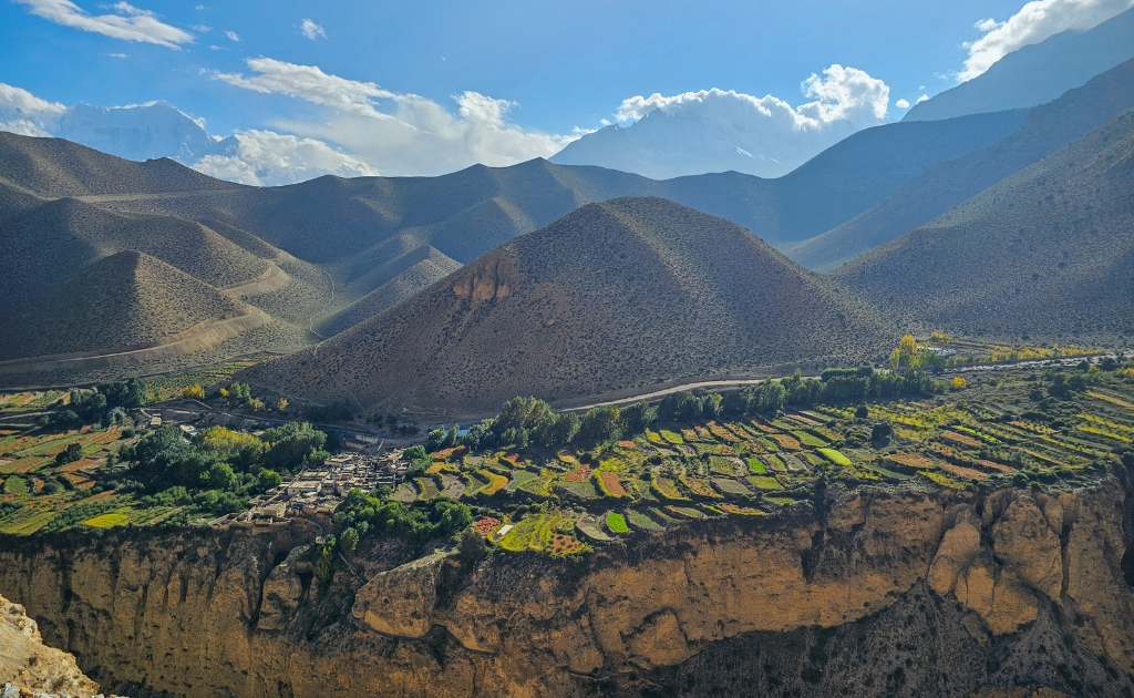 Panoramic view of Upper Mustang's dry Himalayan desert landscape under clear blue skies during the monsoon season, with snow-capped peaks in the background.