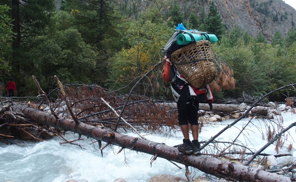 Sherpa porter carrying trekking gear and supplies while crossing a wooden log bridge on the trail toward Rara Lake in western Nepal